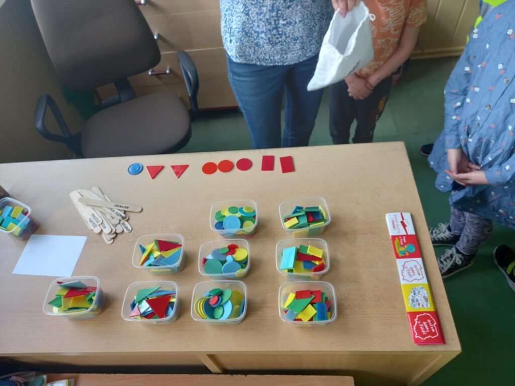 Na biurku stoją pojemniczki z plastikowymi, kolorowymi figurami geometrycznymi
