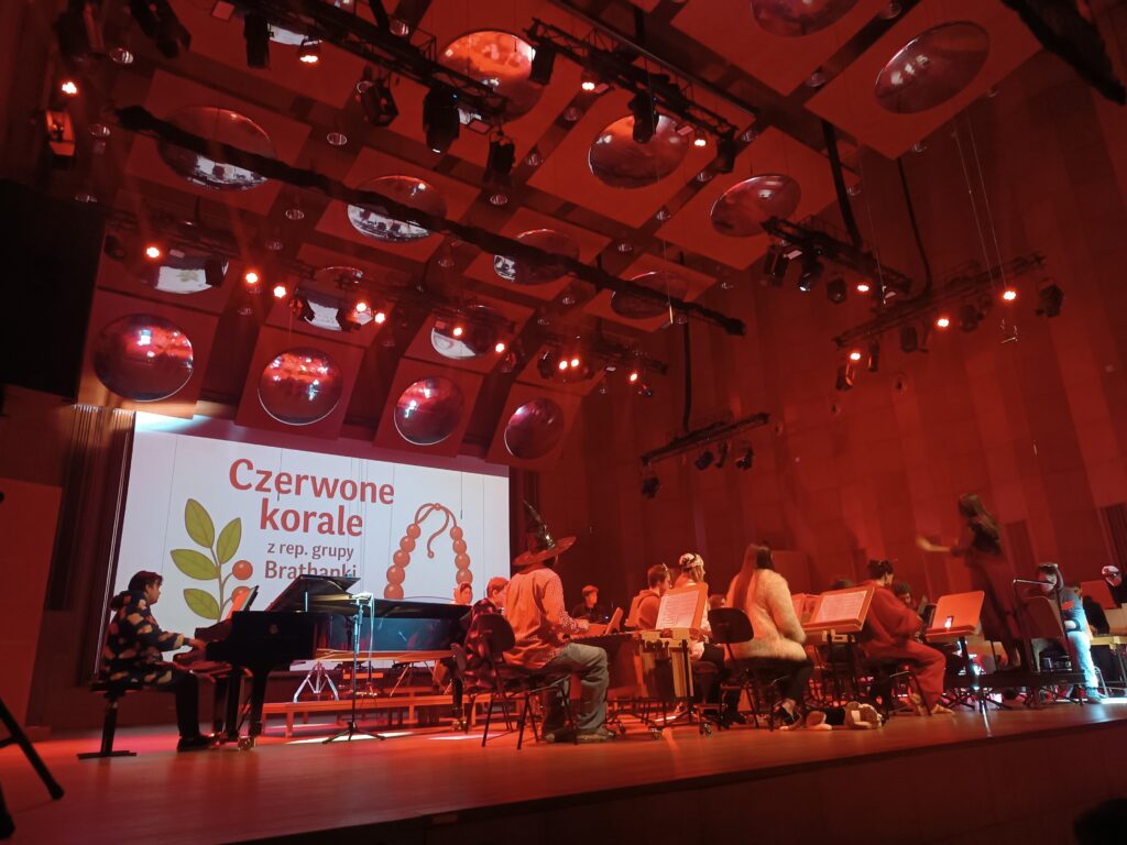 Scena pełna młodzieży grającej na instrumentach. W tle ekran z napisem Czerwone Korale.
