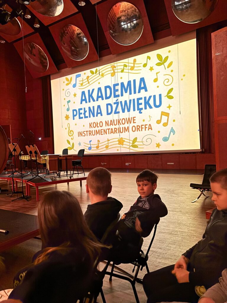 Uczniowie siedzący na krzesłach. W tle wyświetlony ekran z napisem Akademia Pełna Dźwięku.