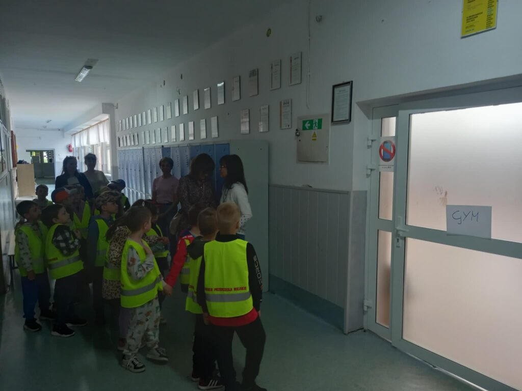 Dzieci w seledynowych kamizelkach stoją na korytarzu z pięcioma paniami