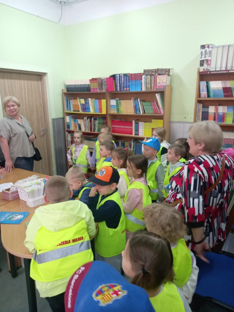 Dzieci w seledynowych kamizelkach stoją w pomieszczeniu z książkami.