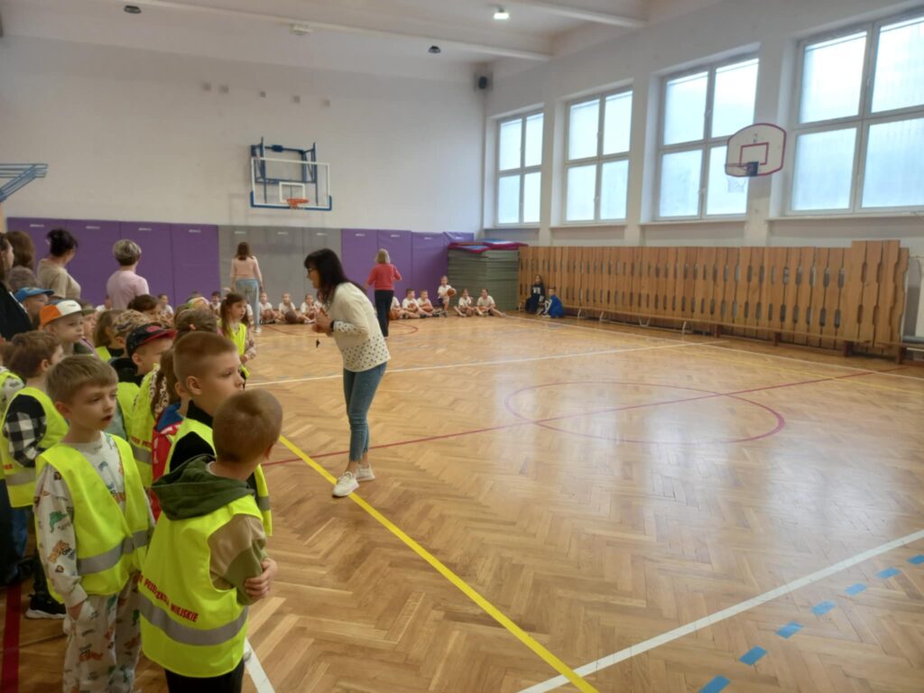 Dzieci w seledynowych kamizelkach stoją na sali gimnastycznej i patrzą na panią, która cos opowiada. W tle pod ścianą siedzą uczniowie w biało czerwonych strojach.