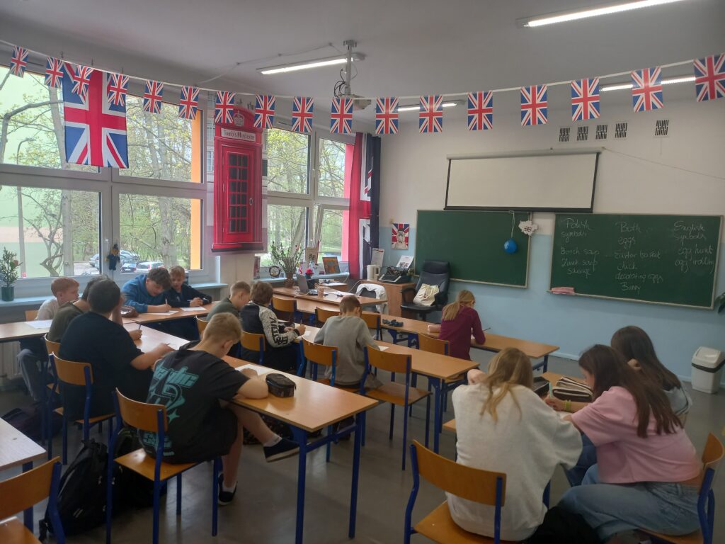 Uczniowie siedzą w ławkach, w tle tablica w pracowni języka angielskiego.