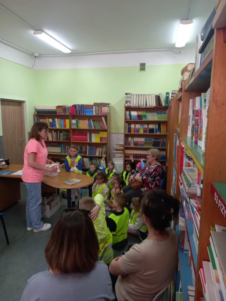 Dzieci w seledynowych kamizelkach siedzą na podłodze i patrzą na panią nauczycielkę, są w pomieszczeniu z książkami, są jeszcze trzy dorosłe panie.