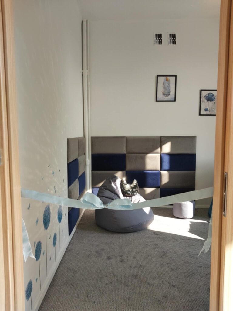 Zdjęcie przedstawia wnętrze pokoju wyciszeni. W tle na ścianie tapicerowane panele i miękka pufa. Na podłodze szara wykładzina. Z przodu widać niebieską wstążkę z kokarą.