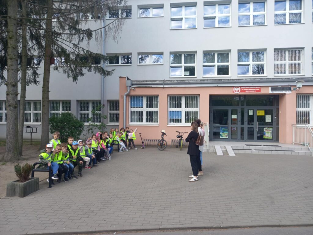 Dzieci w seledynowych kamizelkach siedzą na ławkach przed wejściem do szkoły 182. Stoi trzech opiekunów.