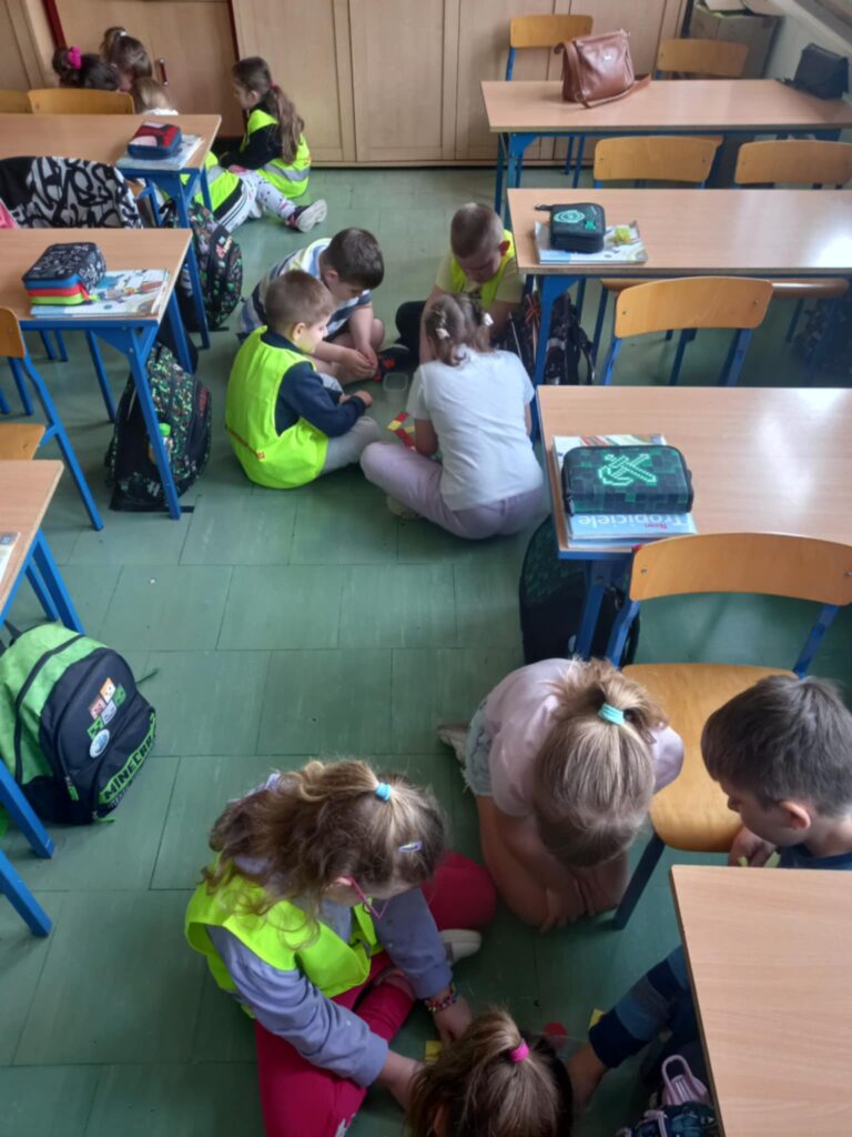 Dzieci w seledynowych kamizelkach i uczniowie podzieleni na czteroosbowe grupy pracują siedząc na podłodze między ławkami.