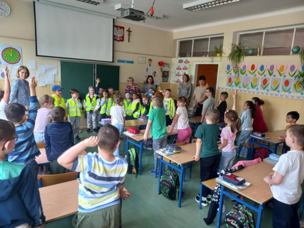 Dzieci w seledynowych kamizelkach i cztery panie stoją w klasie przy tablicy a uczniowie stoją w ławkach. Powitanie.