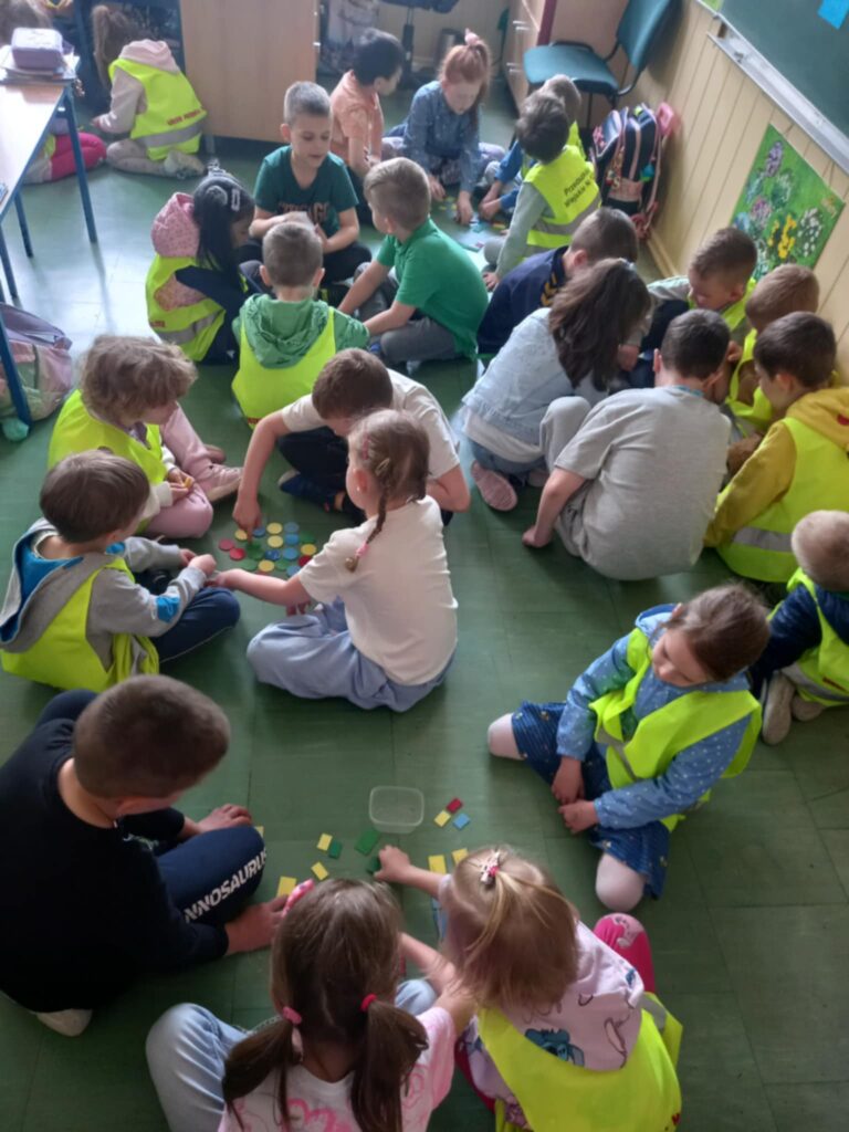 Dzieci w seledynowych kamizelkach i uczniowie podzieleni na czteroosbowe grupy pracują siedząc na podłodze.
