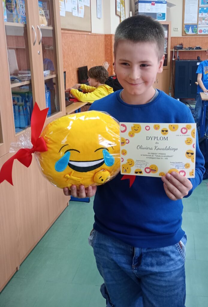 Uczeń z dyplomem i nagrodą, dużą żółtą poduszką w kształcie emotikonki. Na tle klasy.