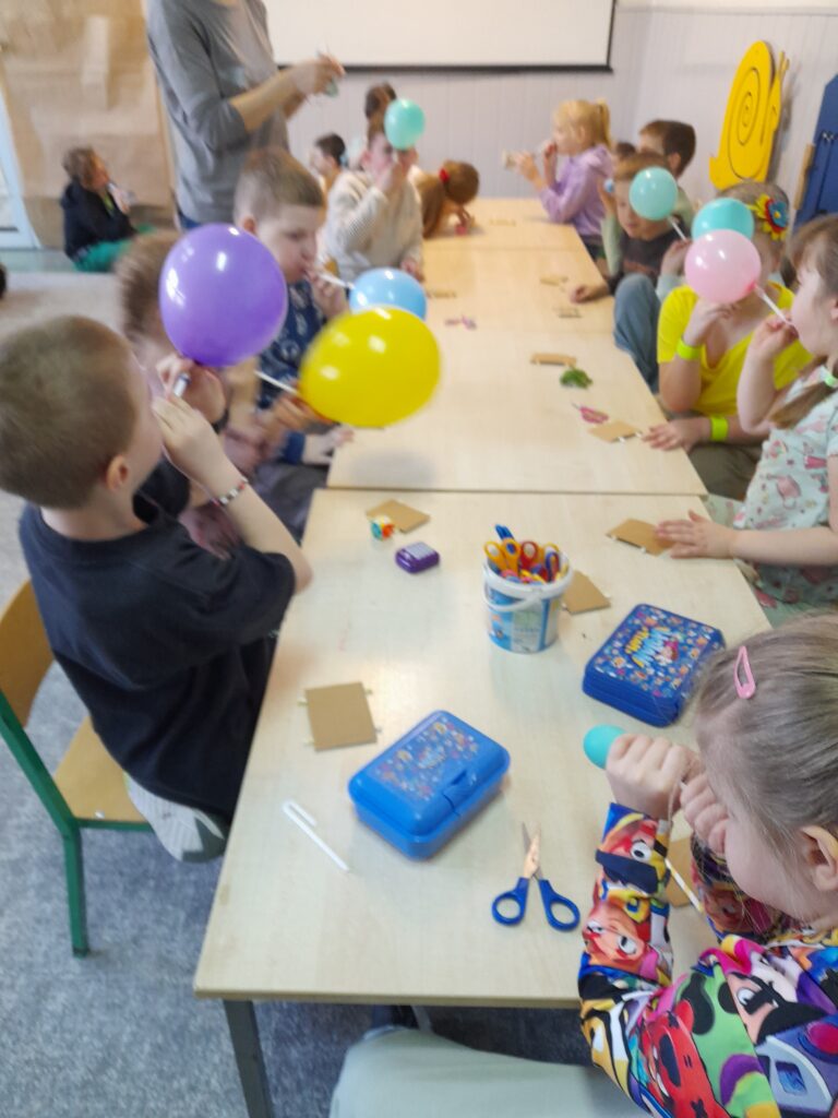 na zdjęciu widzimy jak dzieci siedzą przy stolikach i konstruują pojazdy z balonem.