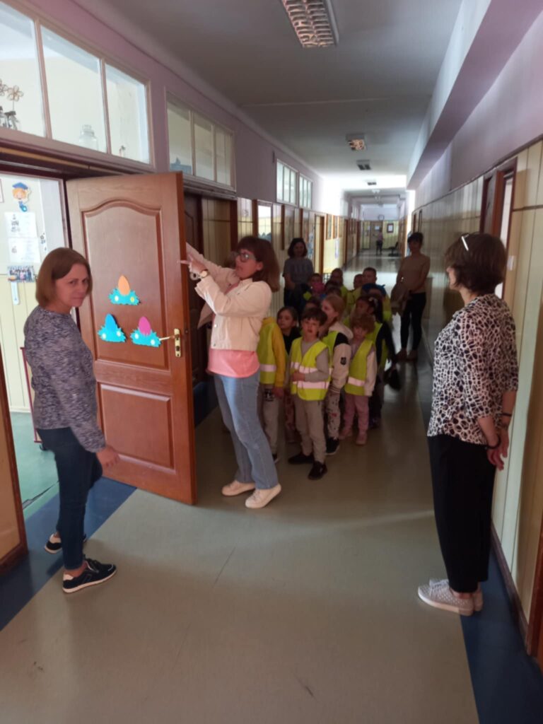 Dzieci w seledynowych kamizelkach stoją na korytarzu przy otwartych drzwiach do klasy. Jest też pięć pań.