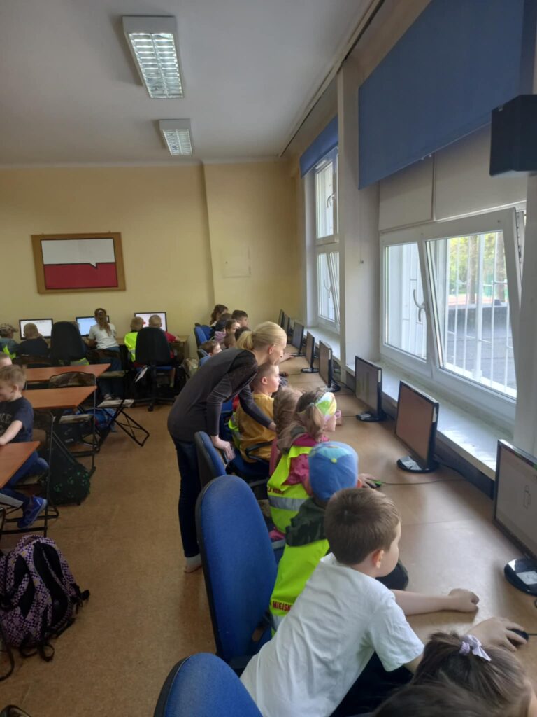 Dzieci w seledynowych kamizelkach i uczniowie siedzą w ławkach przy komputerach, pani nauczycielka pochyla się i tłumaczy zadanie.