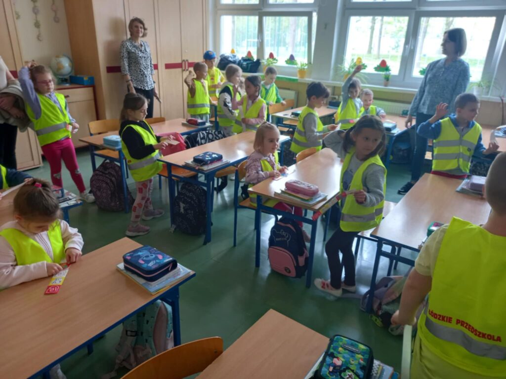 Dzieci w seledynowych kamizelkach siedzą i stoją przy szkolnych ławkach w klasie, w tle nauczycielka.