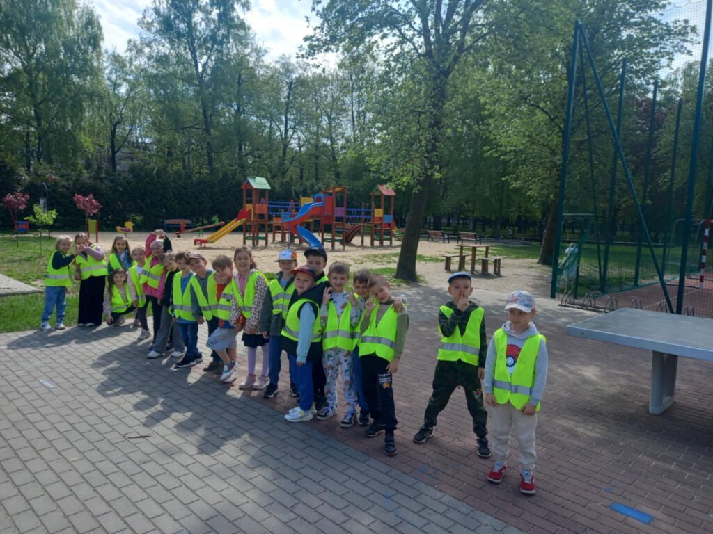 Dzieci w seledynowych kamizelkach stoją w rzędzie, w tle kolorowy plac zabaw