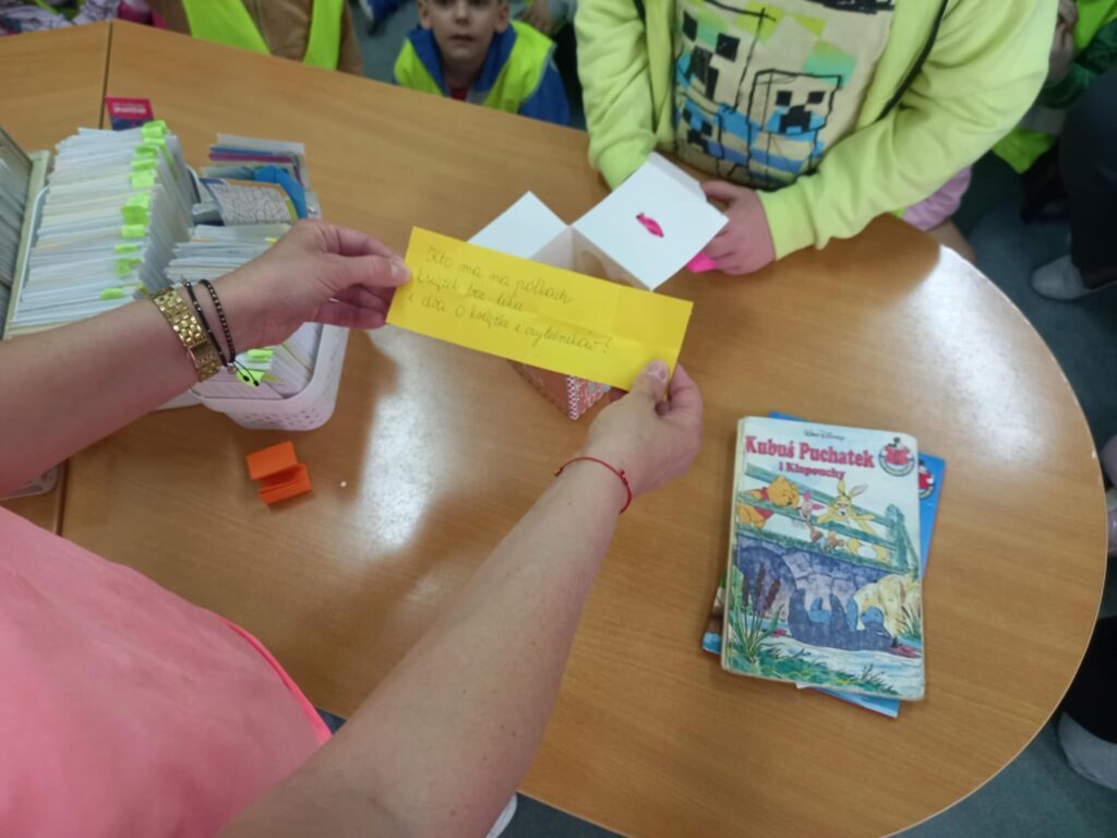 Żółta kartka z zapisaną zagadką: Kto ma na półkach książek bez liku i dba o książki i czytelników? trzymana przez nauczyciela nad biurkiem