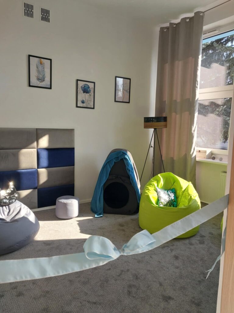Zdjęcie przedstawia wnętrze pokoju wyciszeni. W tle na ścianie tapicerowane panele i miękka pufa. Na podłodze szara wykładzina. Z przodu widać niebieską wstążkę z kokarą.