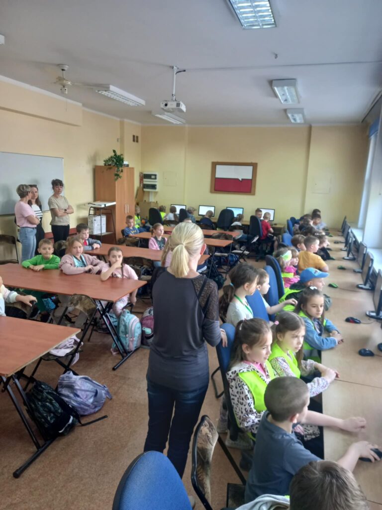 Dzieci w seledynowych kamizelkach i uczniowie siedzą w ławkach przy komputerach, są też cztery panie.