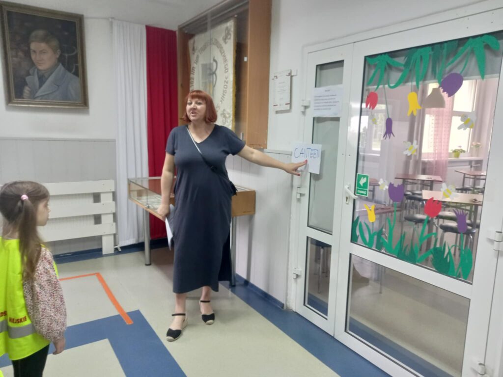 Nauczycielka wskazuje na kartkę z napisem CANTEEN powieszoną na drzwiach
