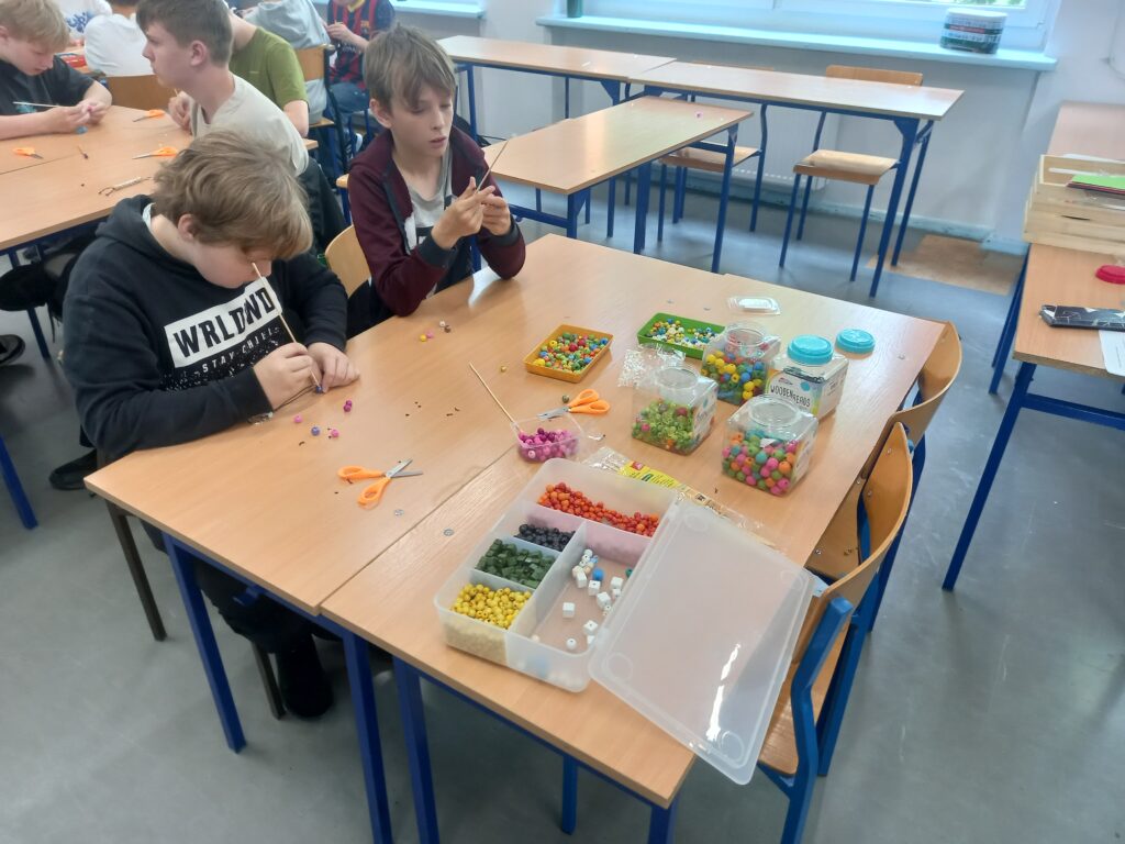 Na ławce znajdują się pudełka z kolorowymi koralikami, obok uczniowie w skupieniu nawlekają wybrane elementy.