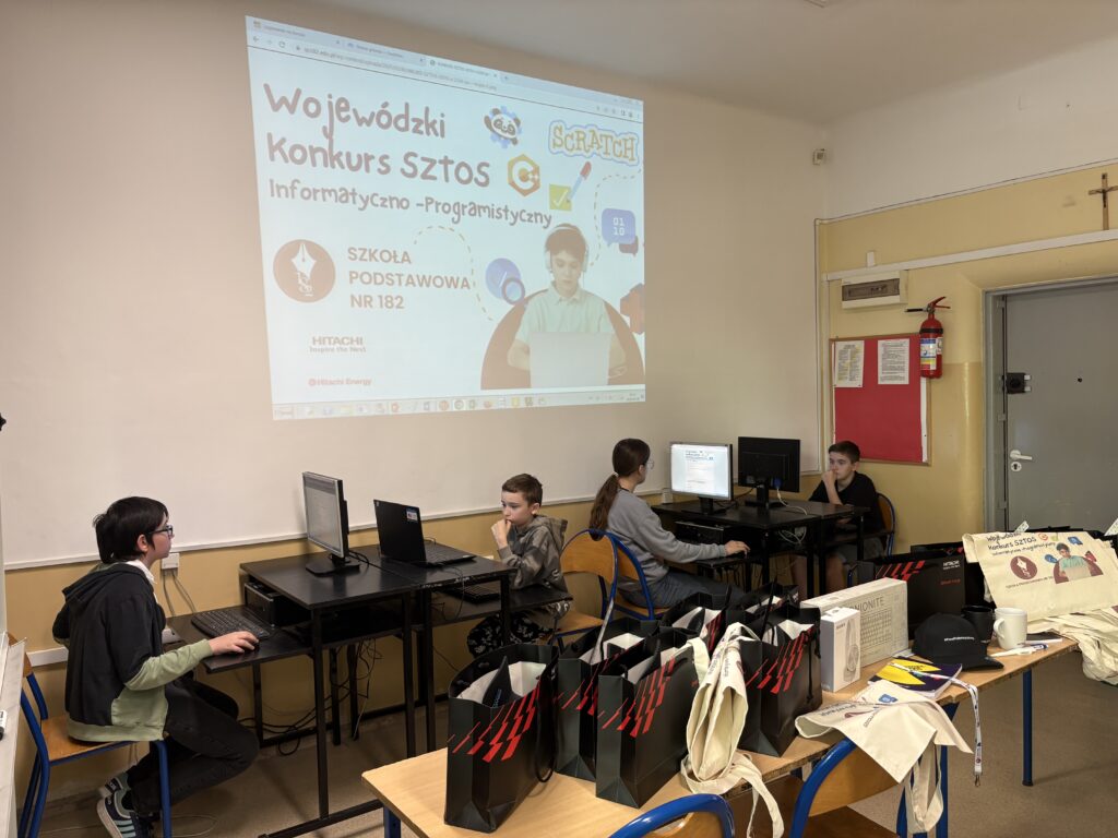 uczniowie siedzą przy komputerze i rozwiązują test, widoczny jest plakat konkursu i nagrody