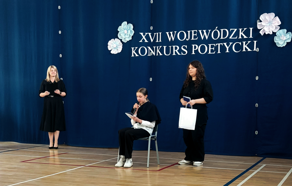 Julia stoi z nagrodą , obok ktoś czyta wiersz
