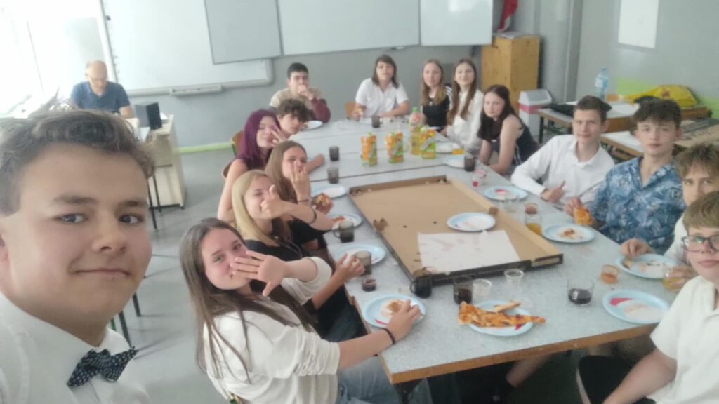 uczniowie siedzą przy stolikach połączonych i jedzą pizzę wspólnie, z boku widać bliżej jednego z uczniów, nauczyciel w oddali
