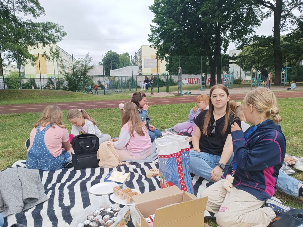 dzieci wraz z panią siedzą na kocykach przed szkołą
