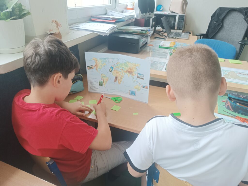 Uczniowie pracują przy biurku uzupełniając kartęy pracy - mapę świata.