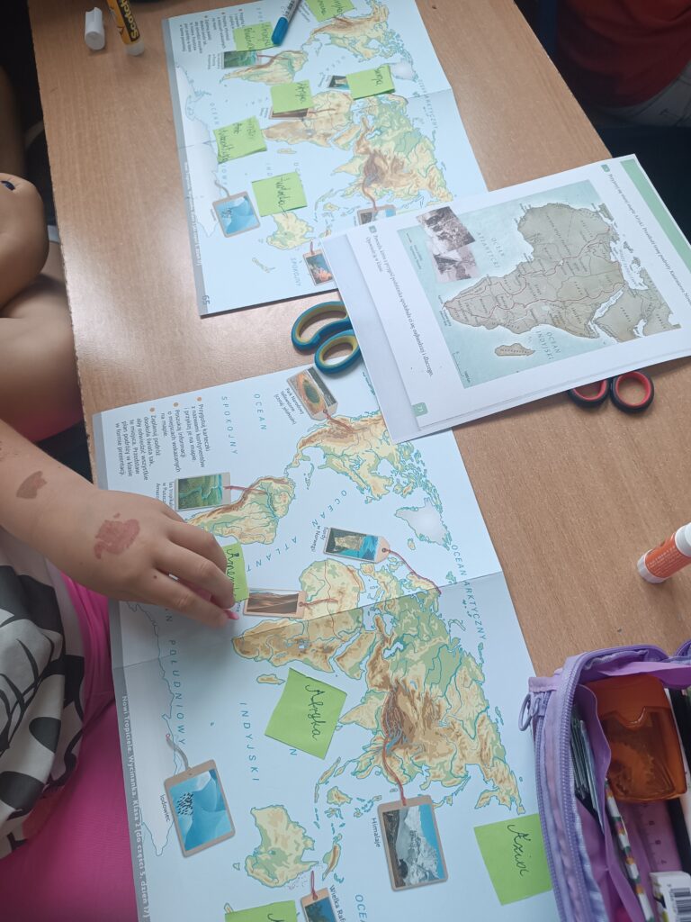 Biurko z rozłożonymi kartami pracy uczniów, mapy świata i mapa Afryki