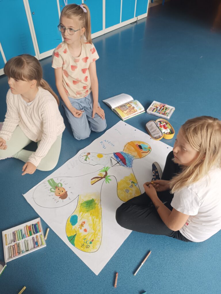 Trzy uczennice siedzą na podłodze i pracują nad dużym kartonem z odrysowanym kształtem człowieka. Dookoła pudełka z kredkami.