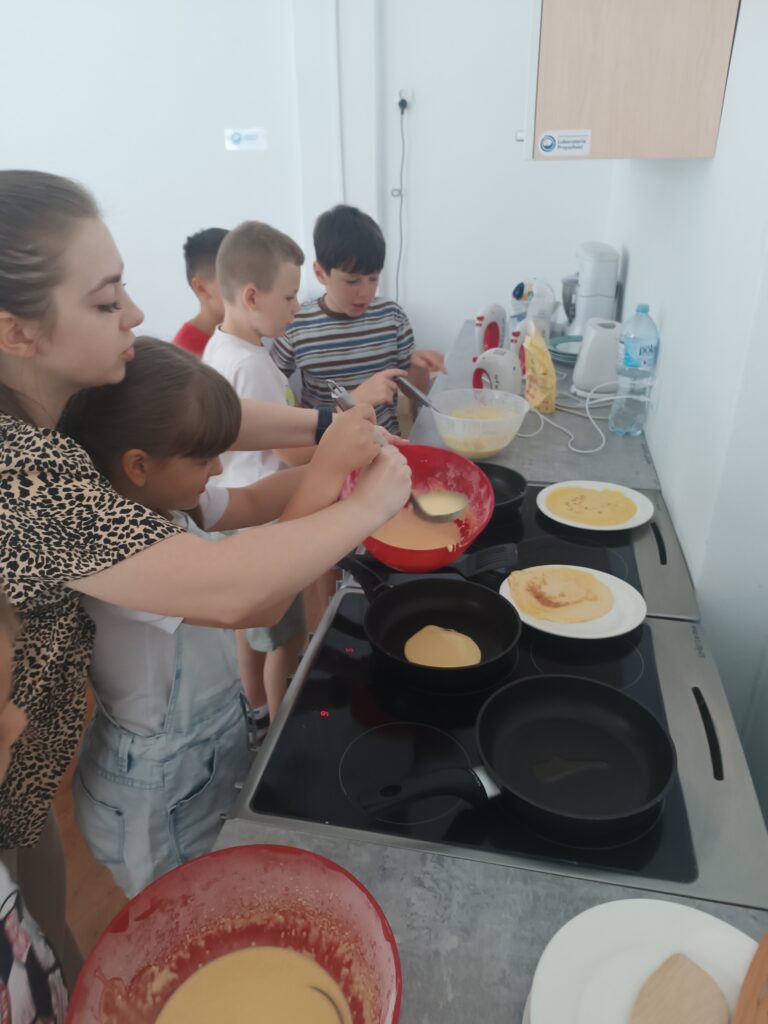 Nauczycielka pomaga uczennicy w nakładaniu ciasta na patelnie. W tle trzech uczniów próbuje nakładać ciasto samodzielnie.