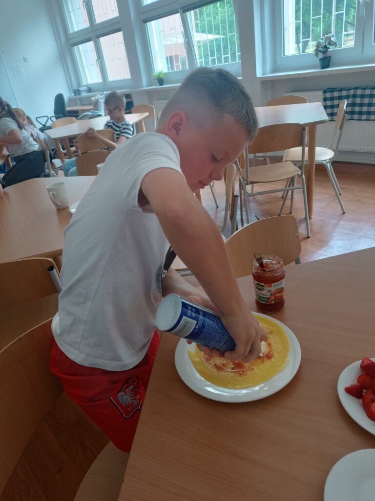 Uczeń przy stoliku nakłada na naleśnika bitą śmietanę z butelki.