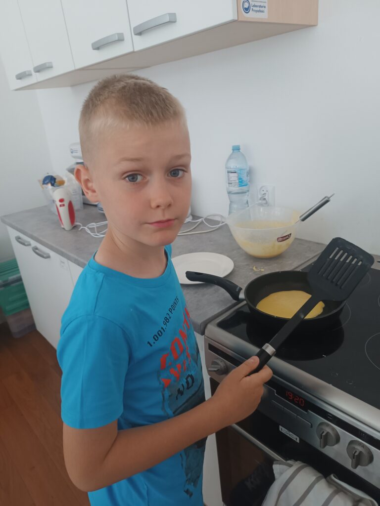 Uczeń stoi z łopatką do naleśników przy kuchence, na palniku patelnia z naleśnikiem.