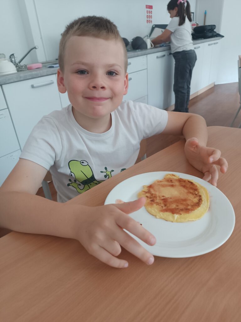 Uczeń z uśmiechem na twarzy siedzi przy stoliku ze swoim naleśnikiem na talerzu.