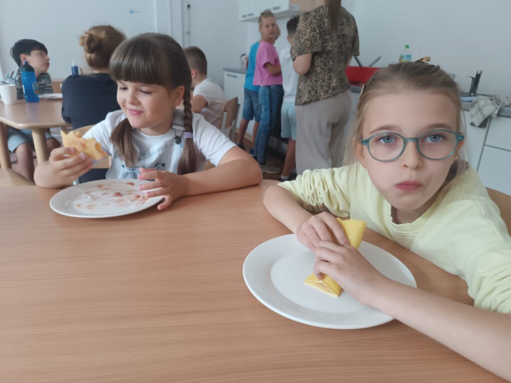 Uczennice z apetytem i uśmiechem jedzą swoje naleśniki rękoma.