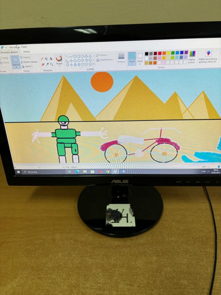 Ekran komputera z pracą ucznia wykonaną w programie paint. Obrazek przedstawia osobę i rower w tle piramidy.