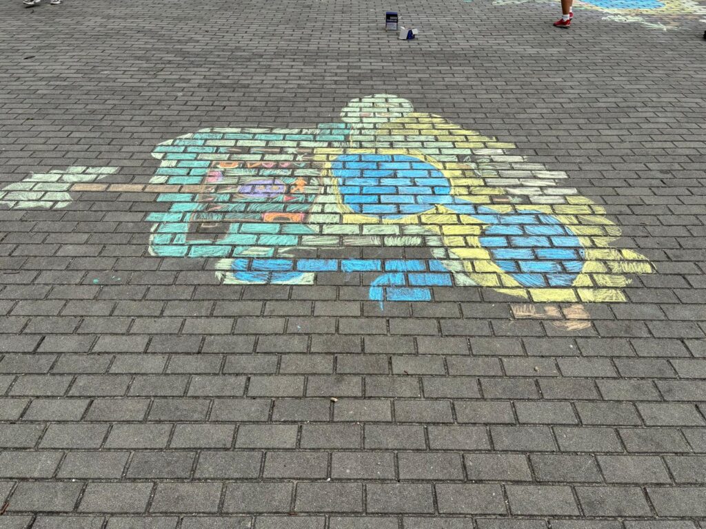 Rysunek na kostce brukowej wykonany kolorową kredą, przedstawia piasek, wodą i palmy.