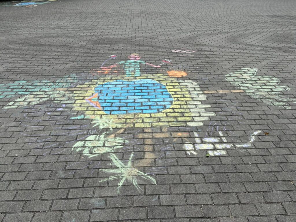Rysunek na kostce brukowej wykonany kolorową kredą, przedstawia postać nad wodą i palmy.