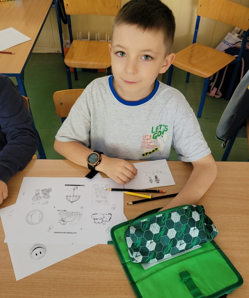 uczeń się uśmiecha, siedzi w ławce, przed nim karty pracy