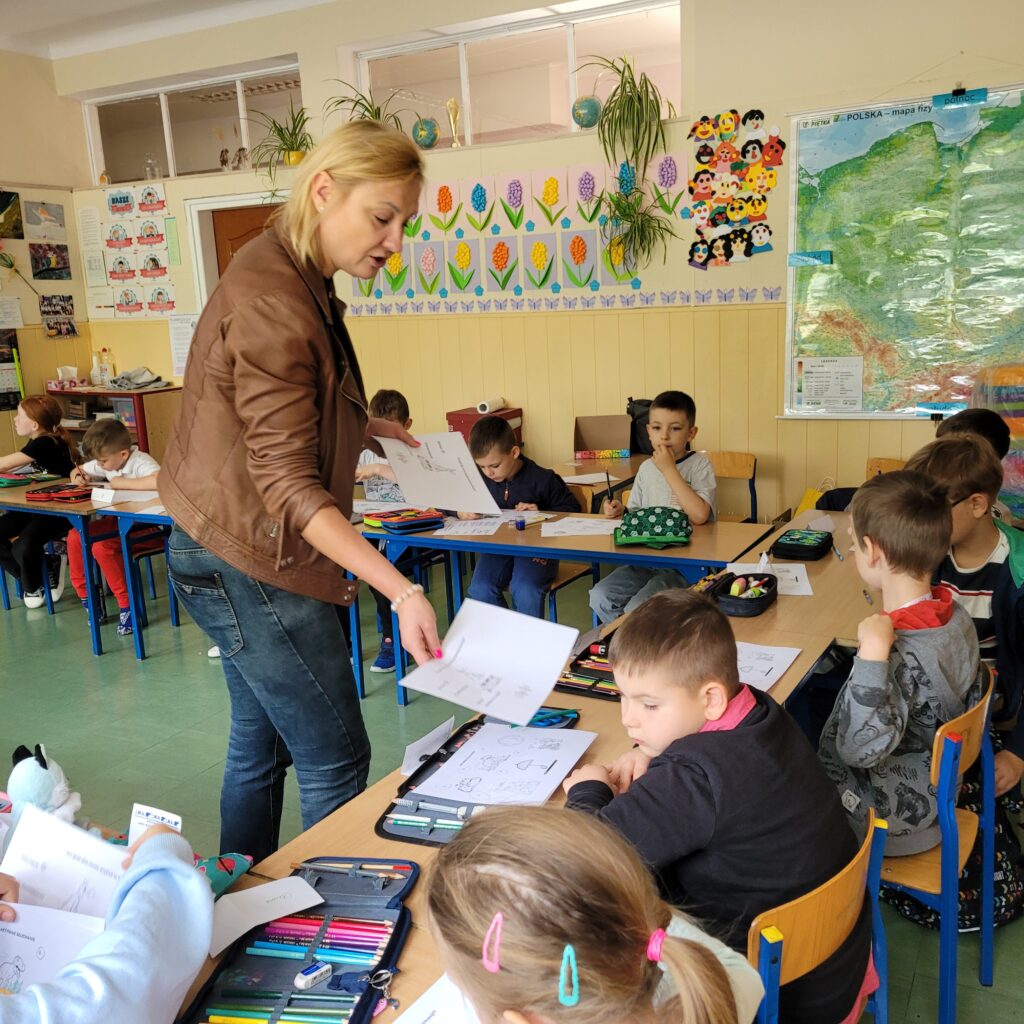 zdjęcie klasy, uczniowie siedzą w ławkach, obok stoi pani i wręcza kartki z zadaniami