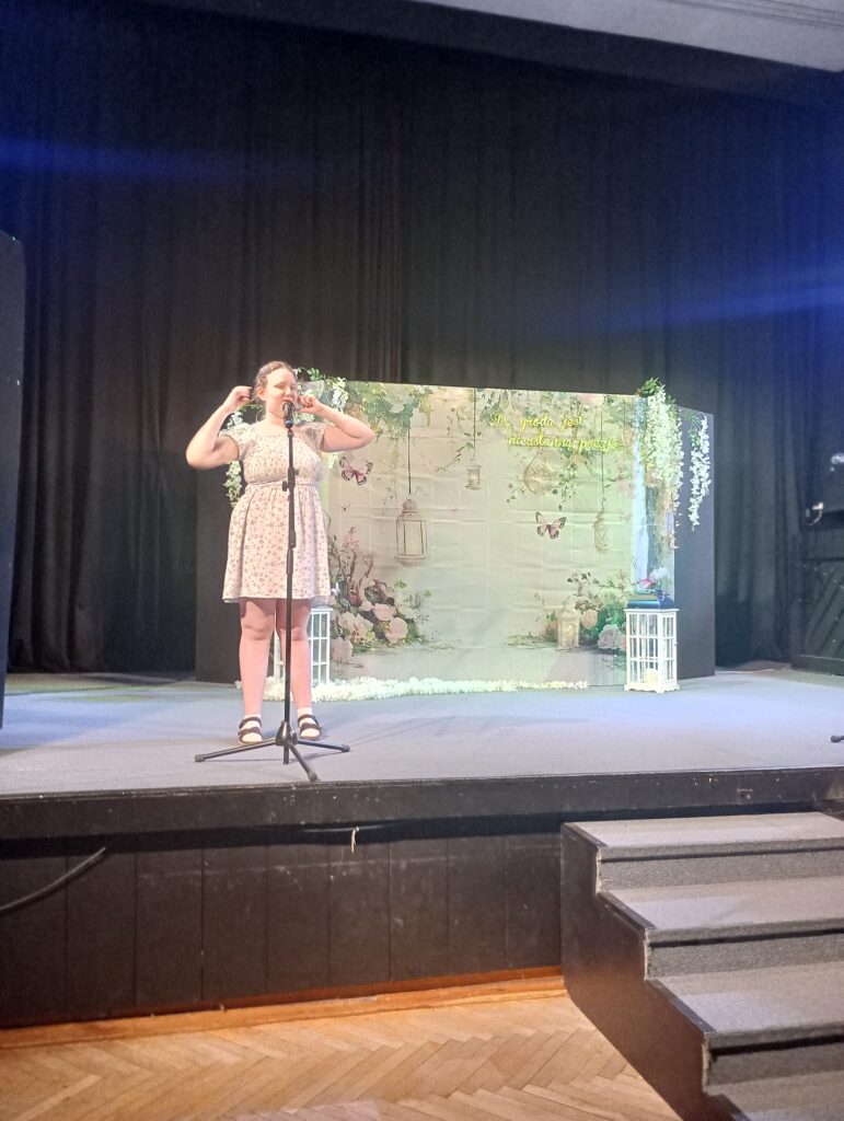 Uczestniczka z mikrofonem podczas recytacji stoi na scenie. Za nią jasne tło z kwiatami i lampiony.