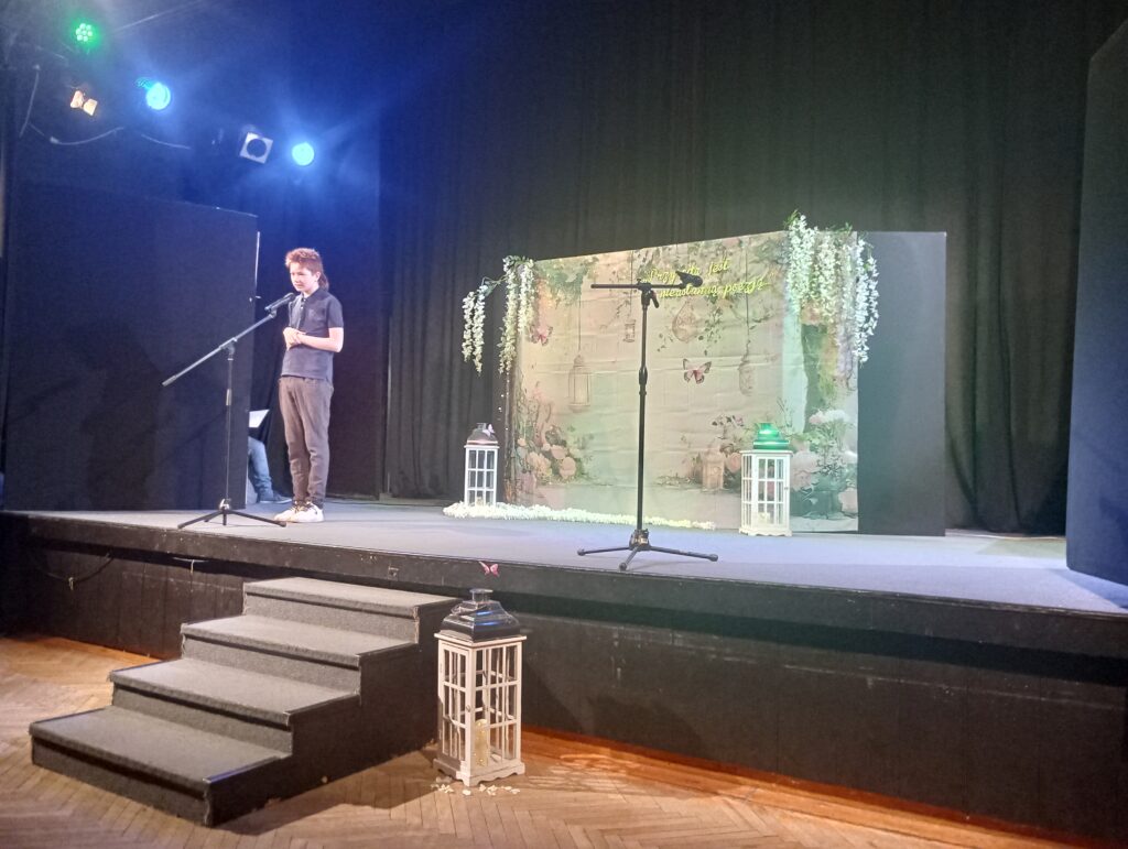 Uczestnik z mikrofonem podczas recytacji stoi na scenie. Za nim jasne tło z kwiatami i lampiony.