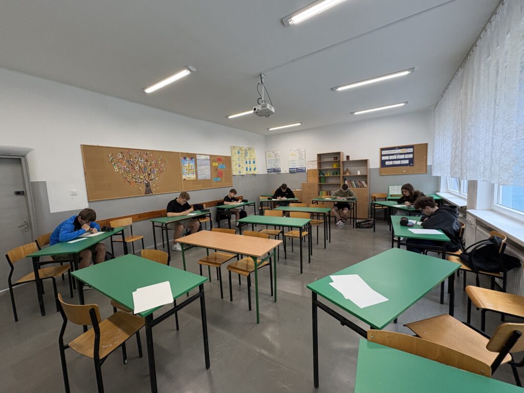 Klasa z ławkami w kolorach brązowym i zielonym. Uczniowie siedzą pojedynczo w ławkach nad kartkami papieru i piszą.