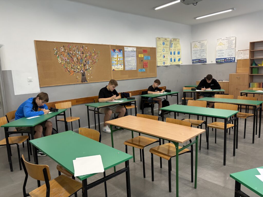 Klasa z ławkami w kolorach brązowym i zielonym. Uczniowie siedzą pojedynczo w ławkach nad kartkami papieru i piszą. Ujęcie zrobione bliżej.