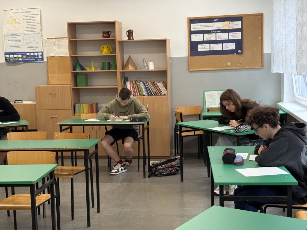 Klasa z ławkami w kolorach brązowym i zielonym. Uczniowie siedzą pojedynczo w ławkach nad kartkami papieru i piszą. Ujęcie zrobione bliżej.