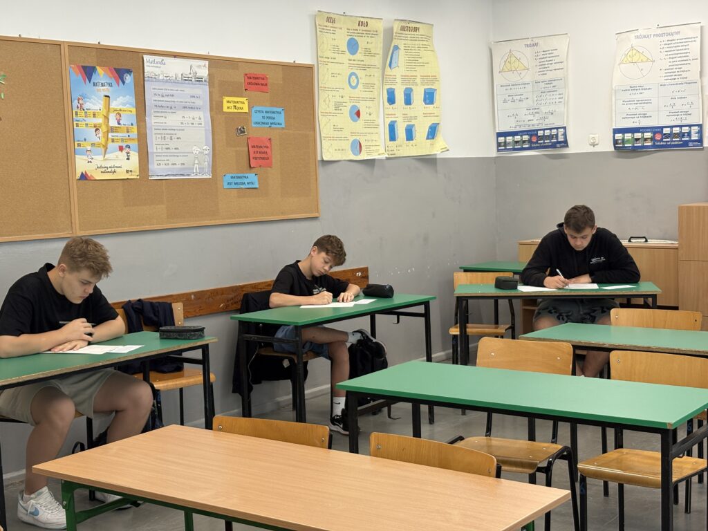 Klasa z ławkami w kolorach brązowym i zielonym. Uczniowie siedzą pojedynczo w ławkach nad kartkami papieru i piszą. Ujęcie zrobione bliżej.