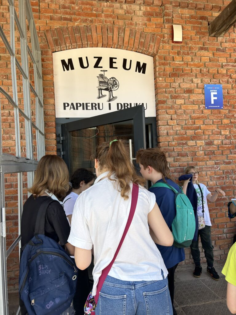 Dzieci stoją przed wejściem do budynku, na którym znajduje się napis: Muzeum Papieru i Druku