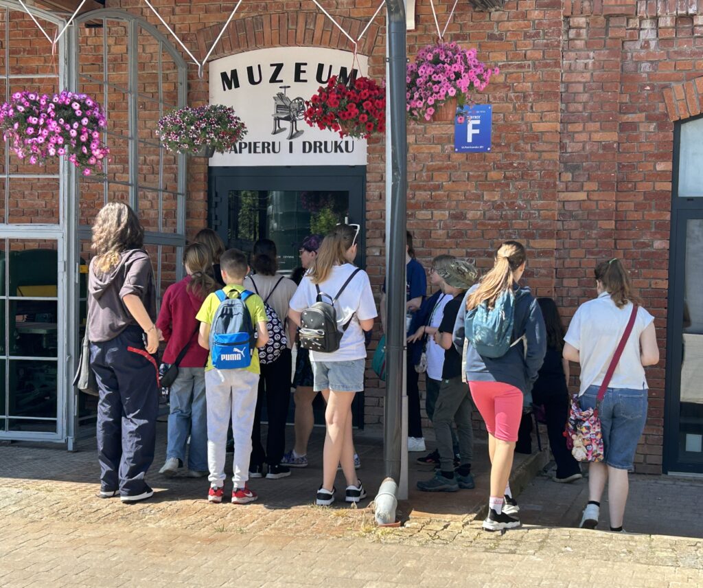 Dzieci stoją przed wejściem do budynku, na którym znajduje się napis: Muzeum Papieru i Druku