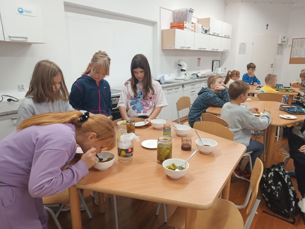 dzieci nakładają sobie produkty do spróbowania, niektórzy siedzą przy stolikach i próbują. 